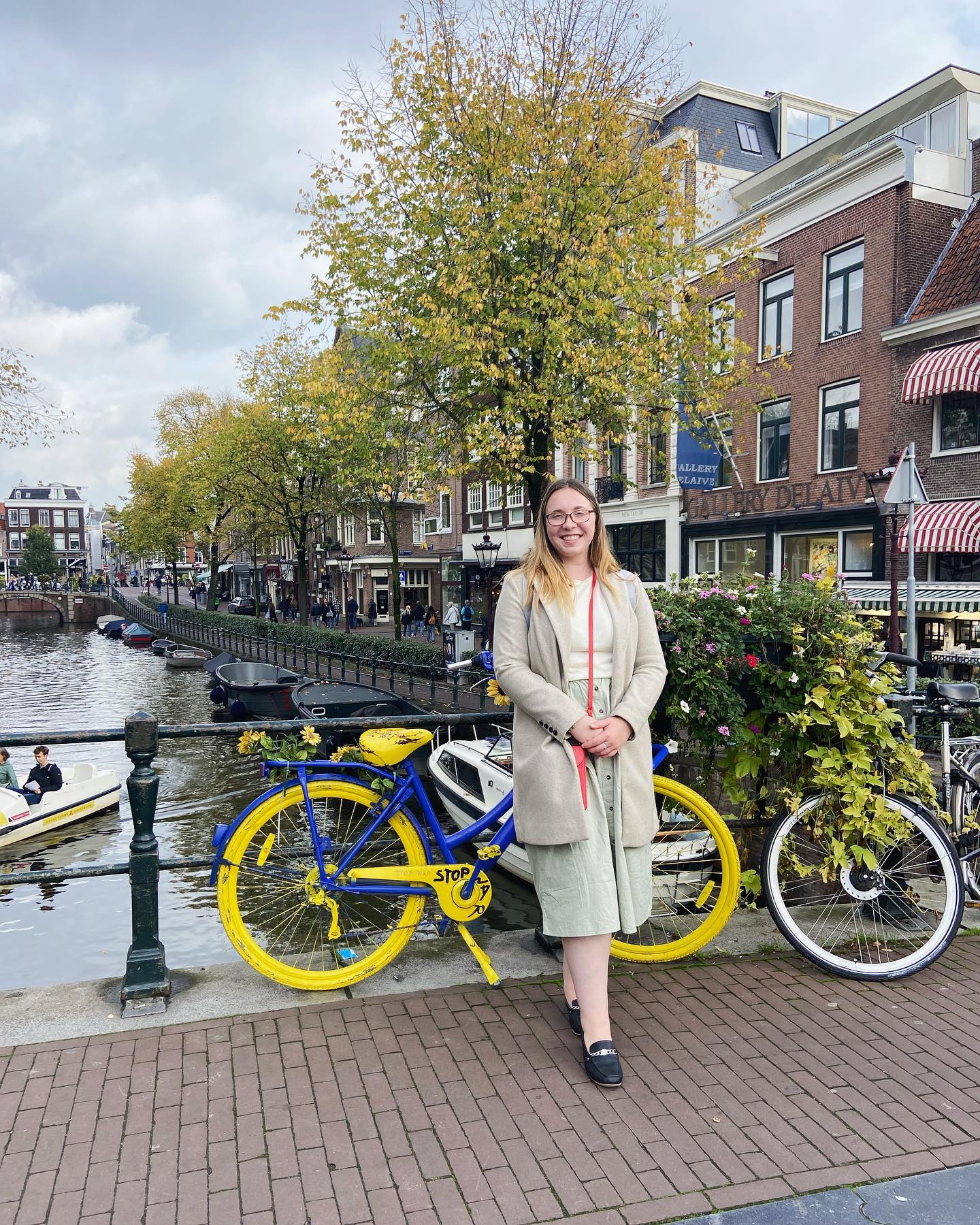 A person stands smiling near a canal in Amsterdam, beside a yellow and blue OV-fiets bicycle