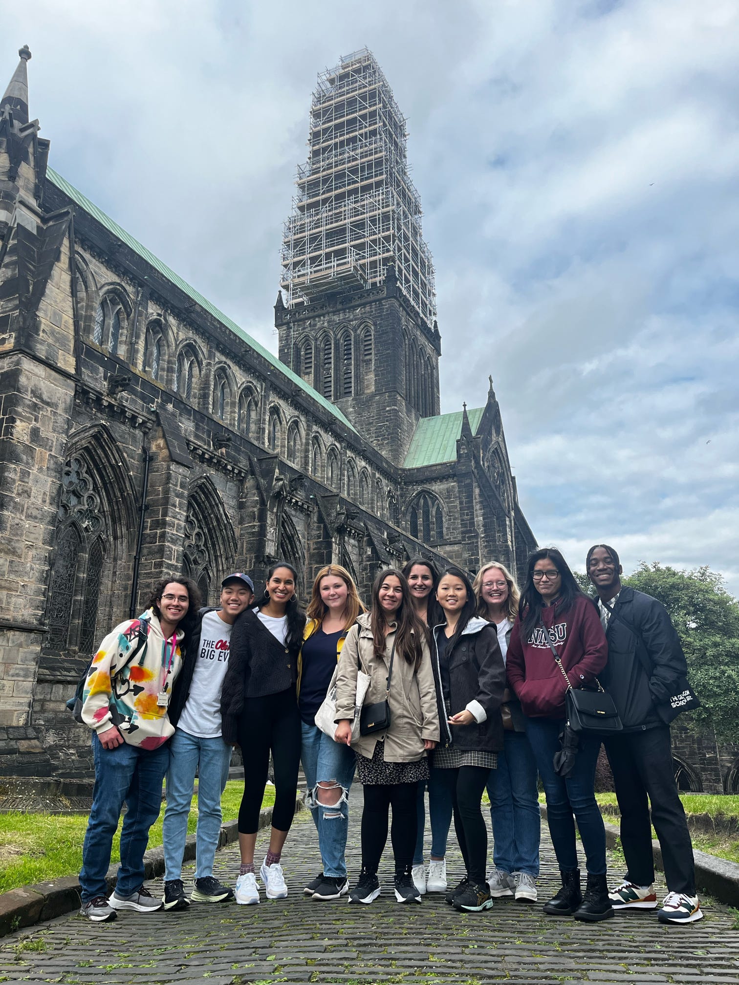 A group of people stands smiling in front of the historic Glasgow Cathedral. The cathedral's tower is partially covered with scaffolding.