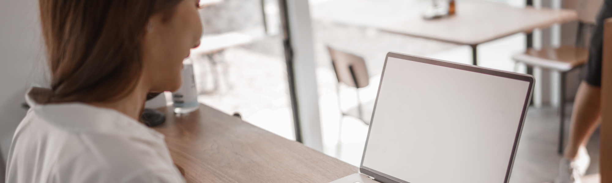A person sits at a wooden table in a modern café, working on a laptop with a blank screen.