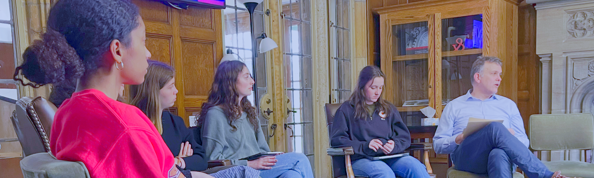 A group of people is seated in a cozy room with wooden panel walls, engaged in a discussion. 