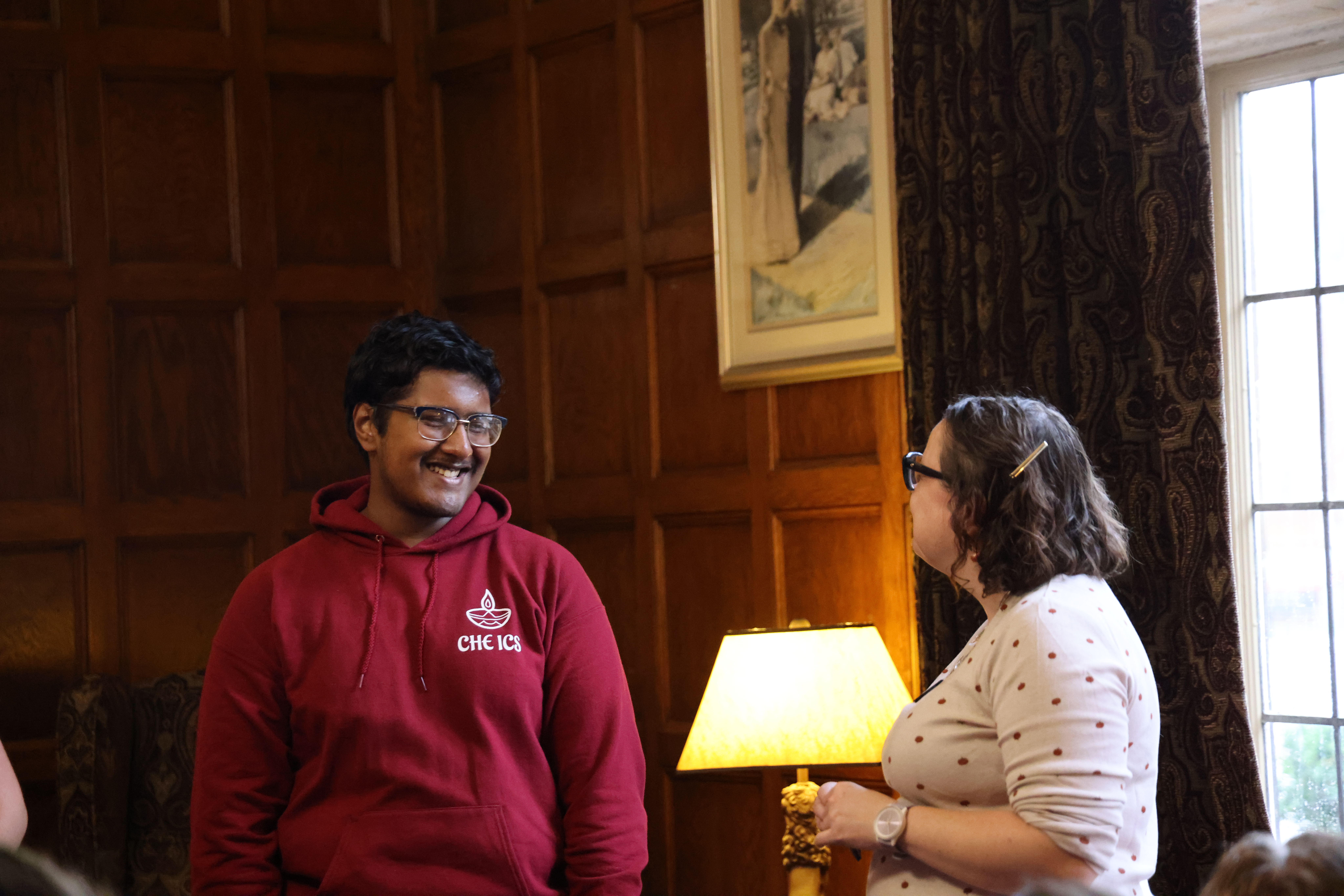 A person in a red hoodie is smiling, standing in a wood-paneled room and another person facing them