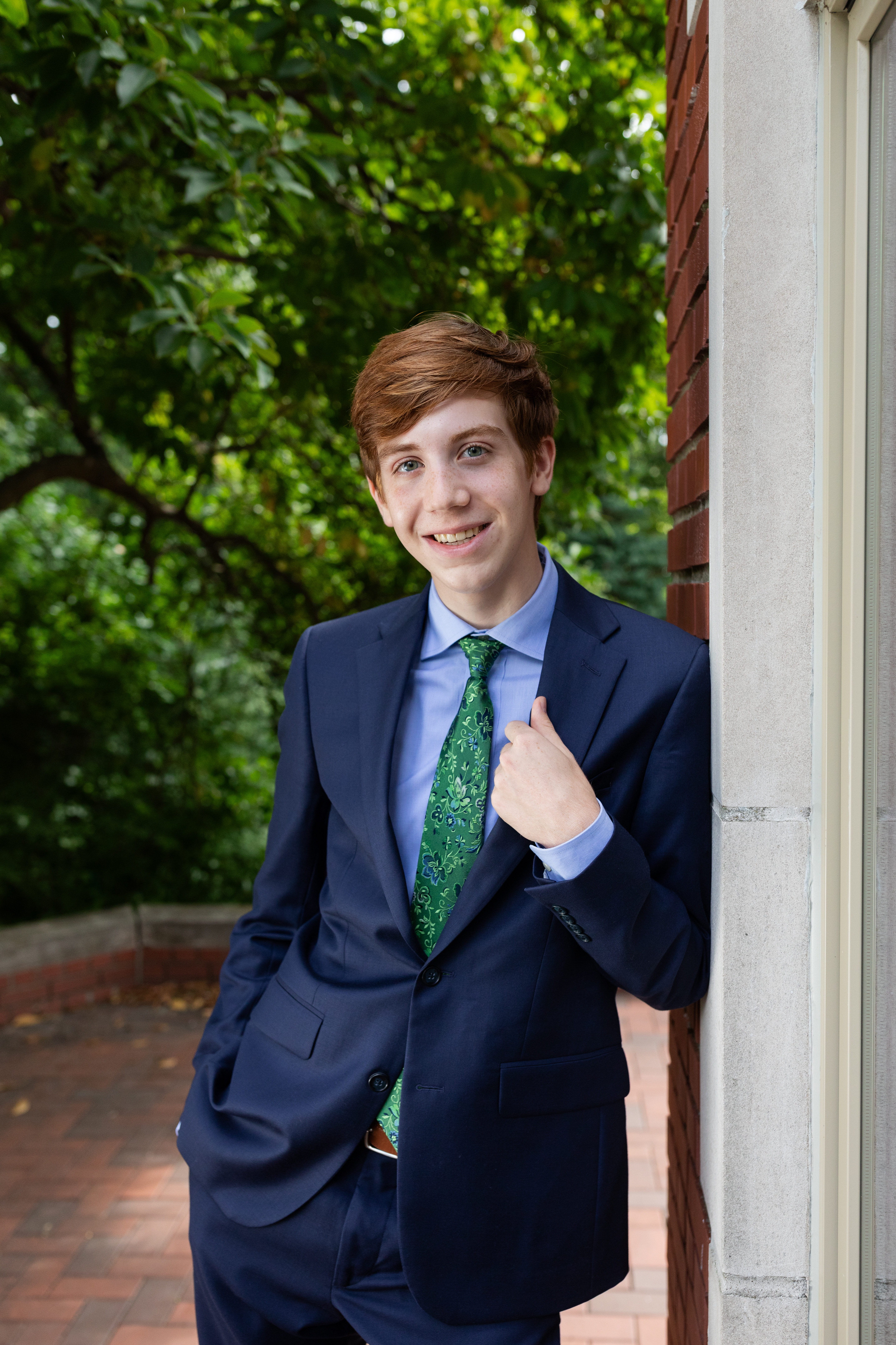 Individual wears blue suit with green tie leaning against brick building with trees in background
