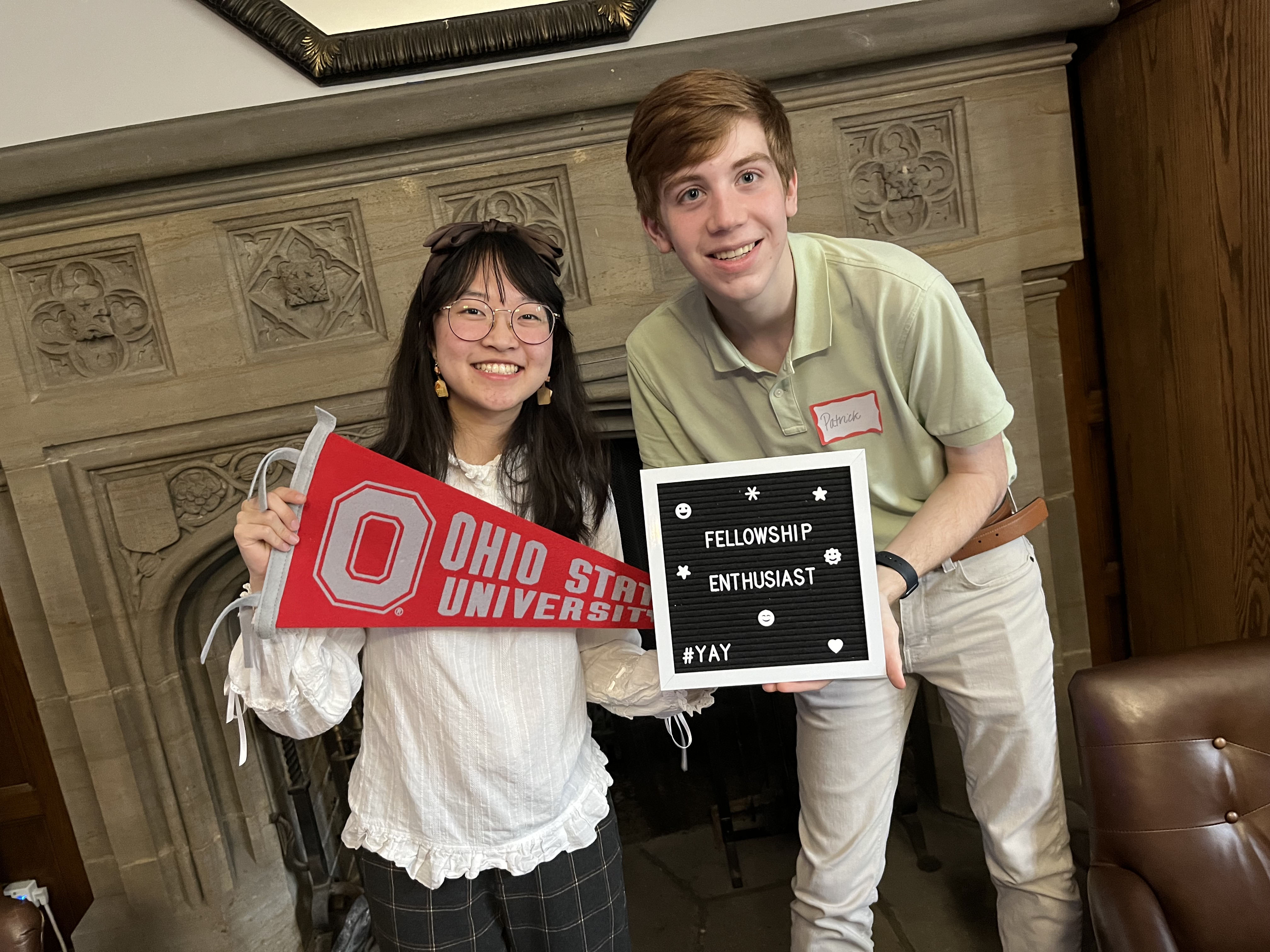 Two people smiling at the camera. One person is holding an Ohio State University pennant. The other holds a letter board