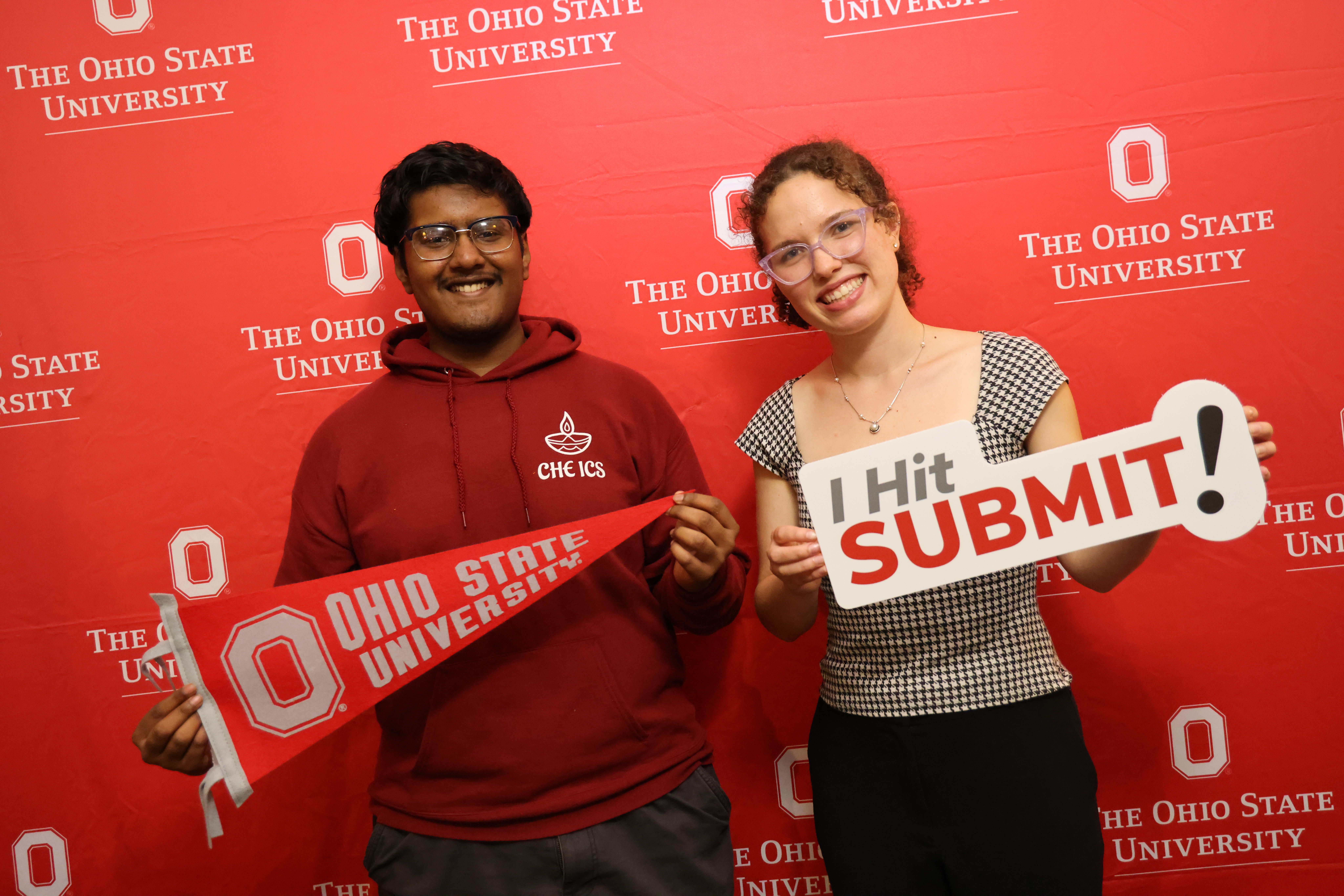 Two individuals smiling in front of a red backdrop holding an Ohio State University pennant and a sign that reads "I Hit SUBMIT!"