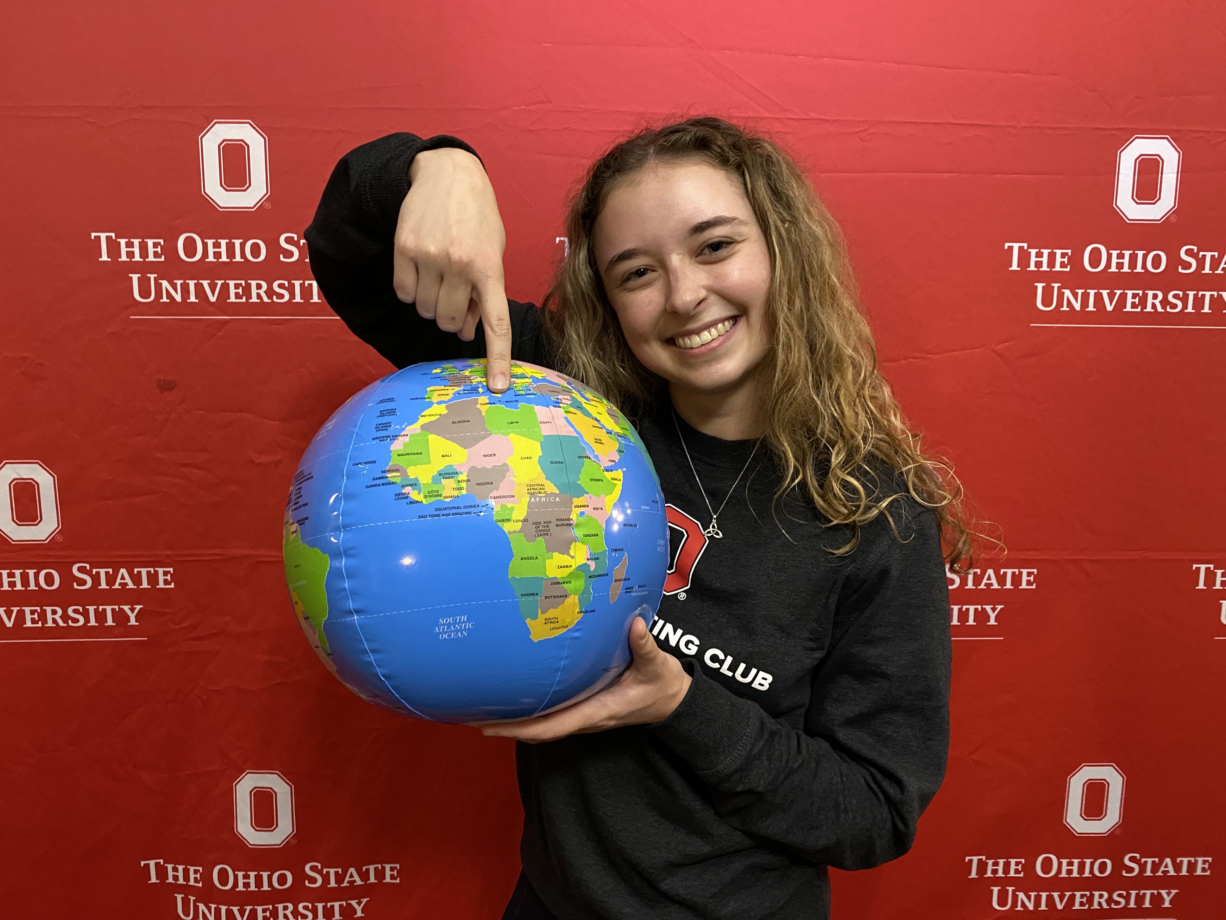 A person stands in front of a red background smiling and holding a small globe, pointing at a location on it.