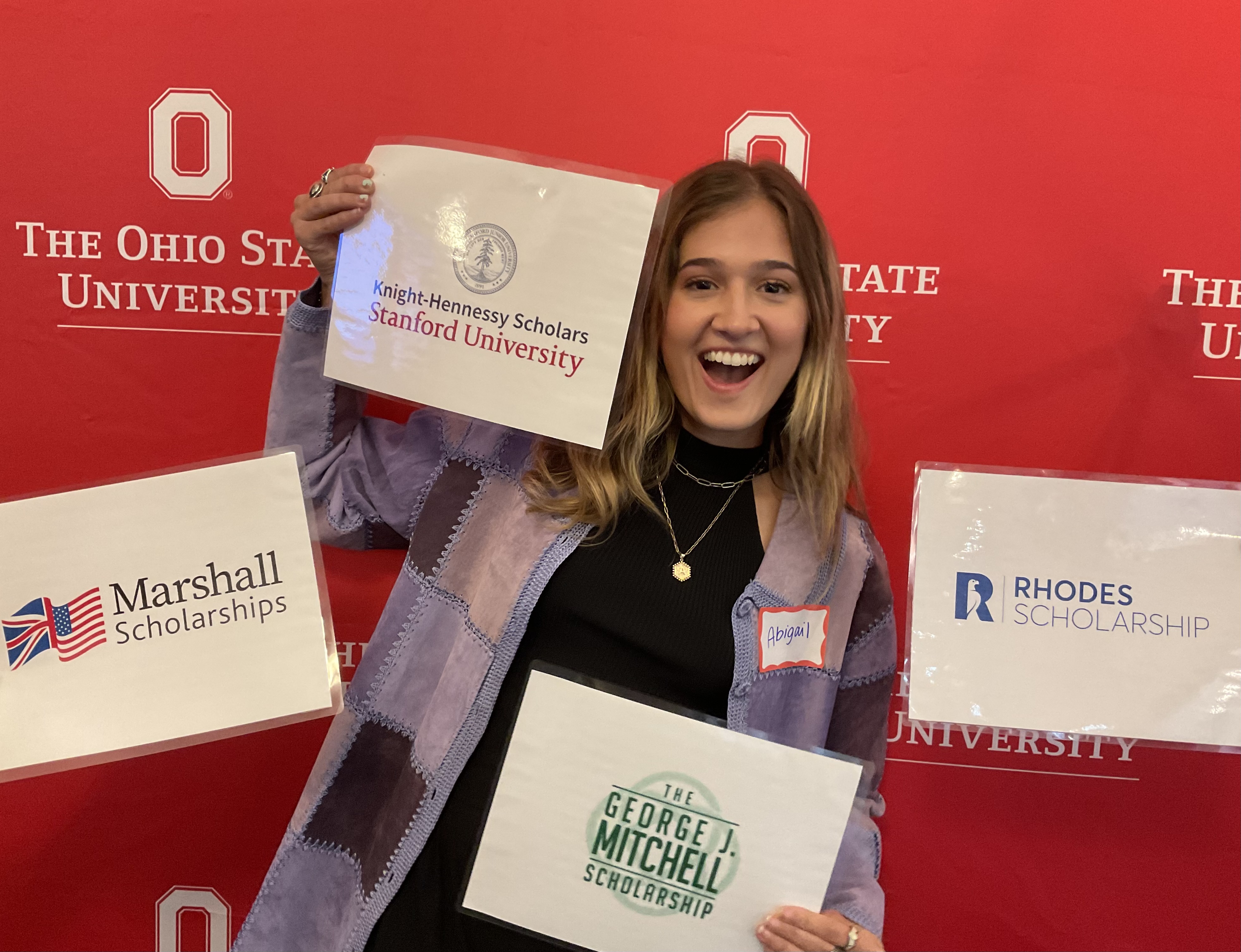 A person smiling standing against a red background surrounded by signs.