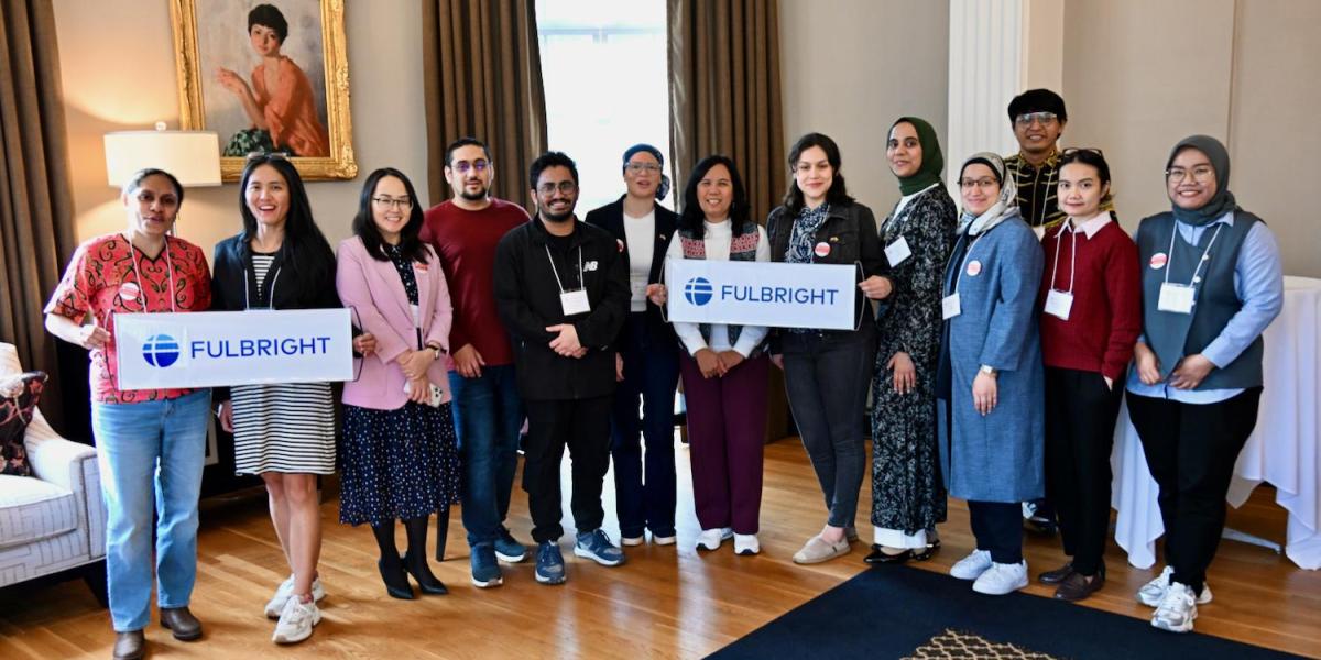 A group of people stands indoors, smiling and holding two signs with the Fulbright logo in a well-furnished room. 