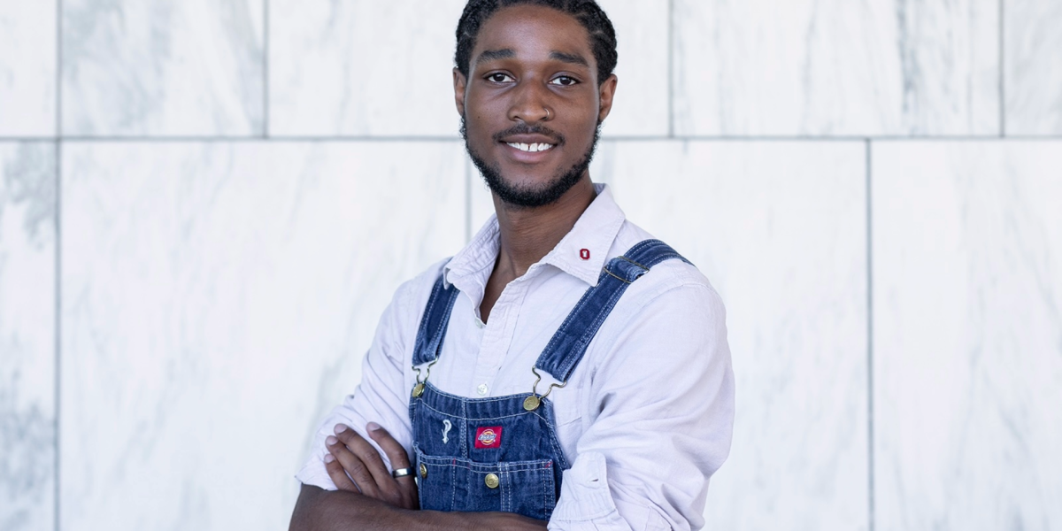 Portrait of D’Laveance Bert-Sims with arms crossed wearing overalls and buttoned up shirt in front of a wall
