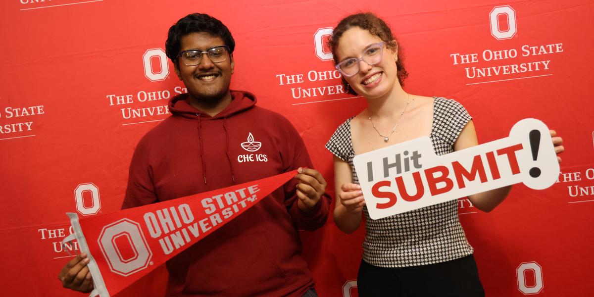 Two individuals smiling in front of a red backdrop holding an Ohio State University pennant and a sign that reads "I Hit SUBMIT!"