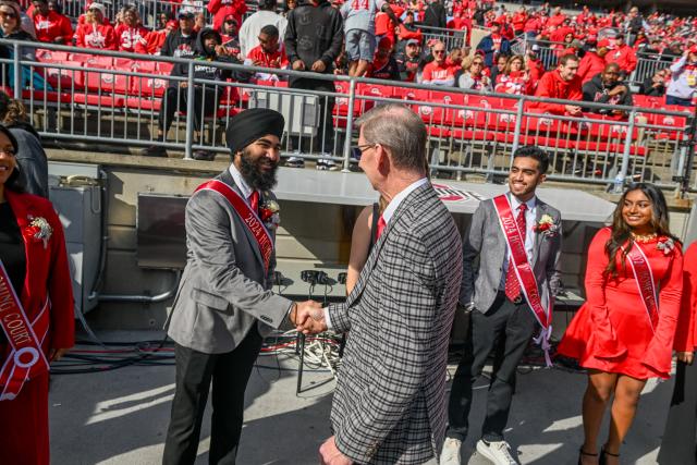 A group of individuals wearing red and white sashes stands on a football field. They are being greeted by a person in a checkered suit. 