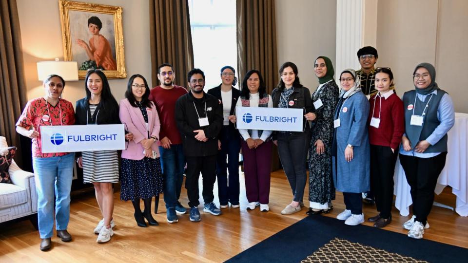 A group of people stands indoors, smiling and holding two signs with the Fulbright logo in a well-furnished room.