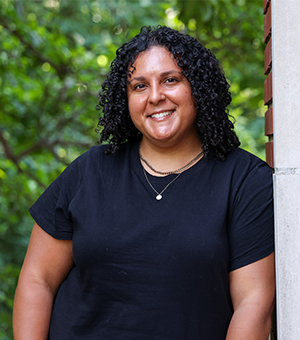 Courtney Gandy smiles, with curly hair, standing outdoors against a wall with a leafy background.