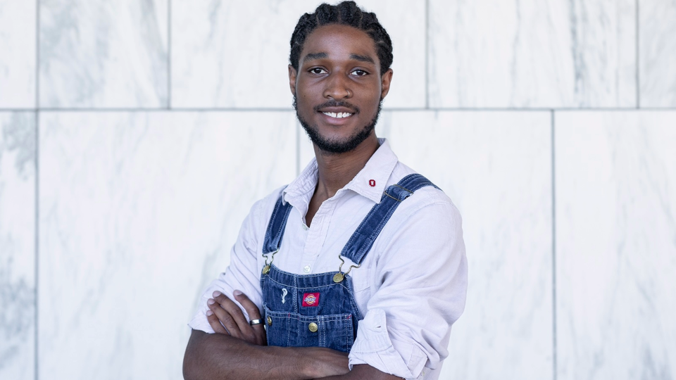 Portrait of D’Laveance Bert-Sims with arms crossed wearing overalls and buttoned up shirt in front of a wall