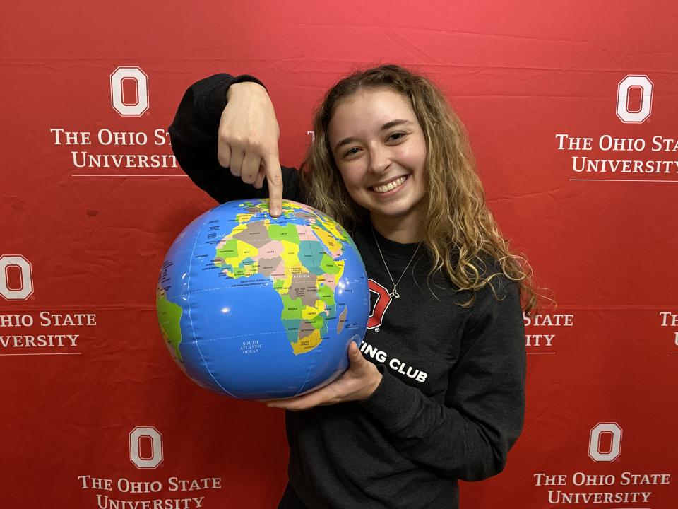 A person stands in front of a red background smiling and holding a small globe, pointing at a location on it.